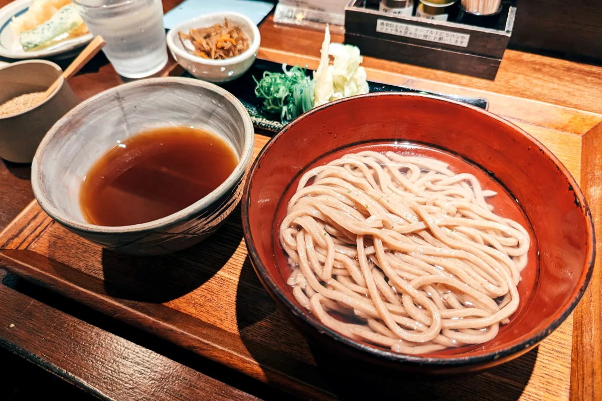 A bowl of cold pale noodles beside a cup of dark dipping sauce