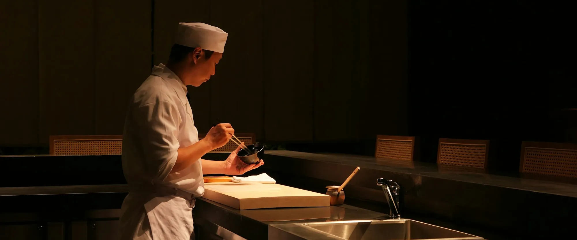 A sushi chef plating at a dark restaurant counter under warm light
