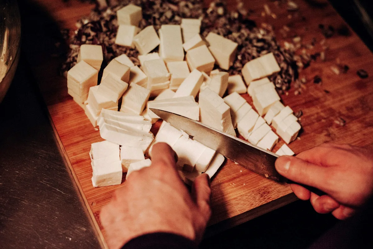 A knife cutting a block of tofu into cubes on a wooden board