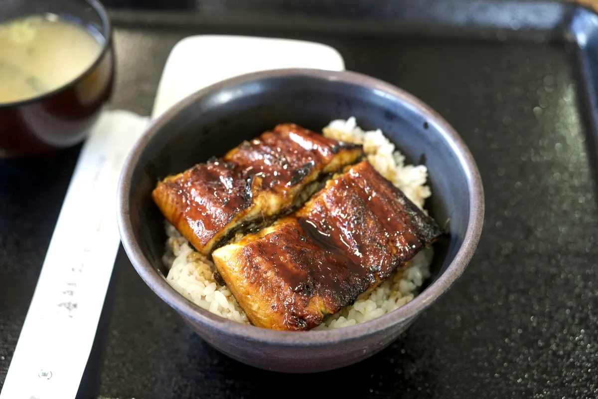 A bowl of rice topped with glazed grilled eel and a bowl of soup alongside