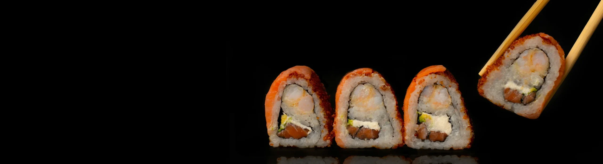 Four salmon-topped sushi rolls on a black reflective surface, one lifted with chopsticks