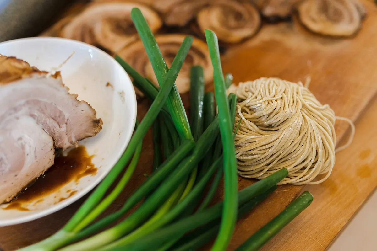 Sliced braised pork, whole green onions and coiled fresh noodles on a wooden board