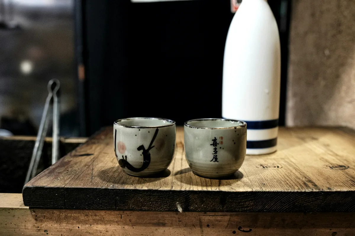 A white sake bottle and two small ceramic cups on a wooden board