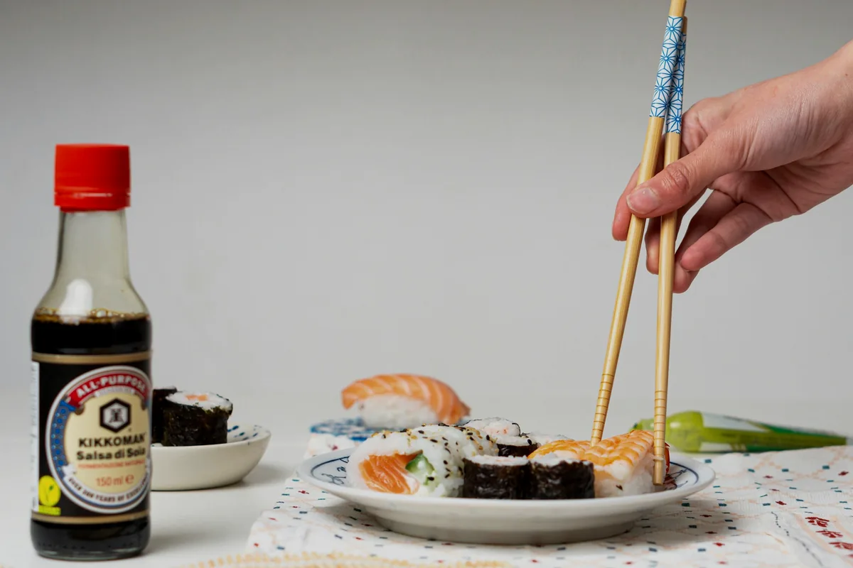 A bottle of soy sauce on a table beside a hand lifting food with chopsticks