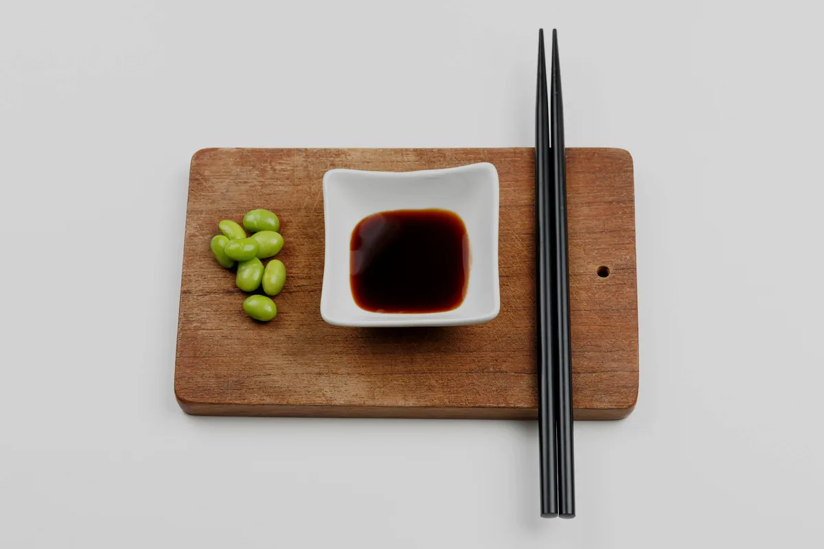 A small dish of soy sauce and chopsticks on a wooden board with edamame