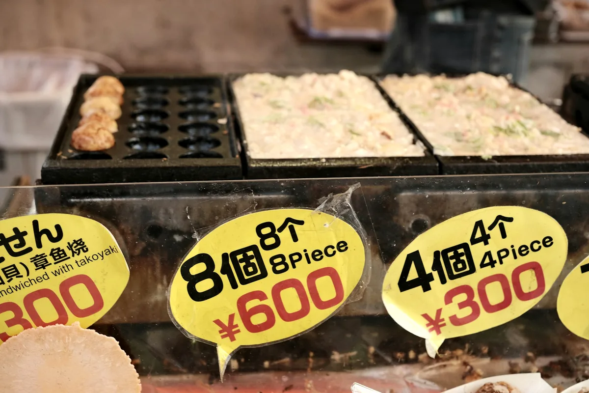 A takoyaki griddle at a stall with price signs above it