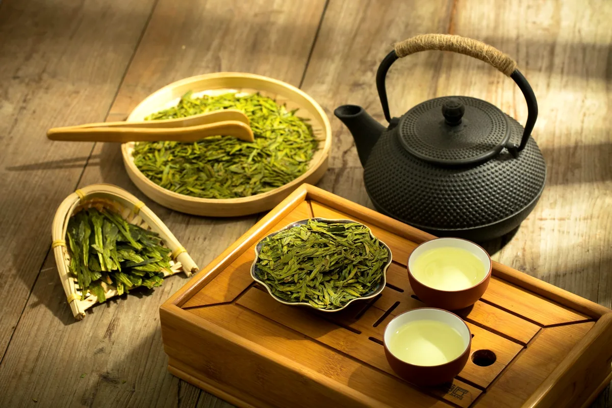 A cast-iron teapot on a wooden tray beside bowls of loose green tea leaves