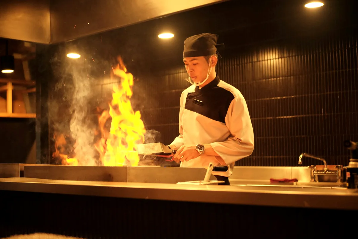 A cook working over a flat griddle as flames rise from the pan
