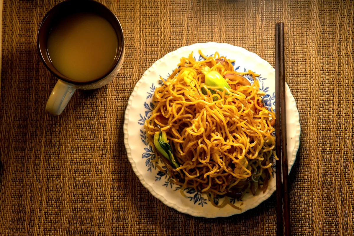 An overhead view of pan-fried yellow noodles on a plate beside a cup of tea